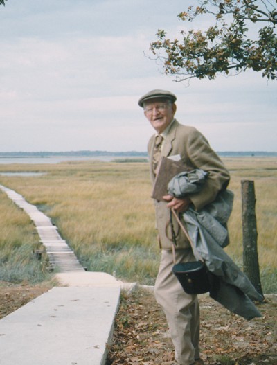 A photo from the 1950s with an older man in a cap and glasses holding binoculars in a case and a jacket standing next to a wooden boardwalk that goes off in the distance toward a marsh. 