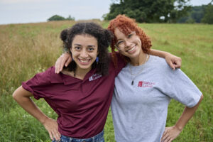 Two young women of color stand in Natural Lands logo shirts with their arms around each other and smile at the camera.
