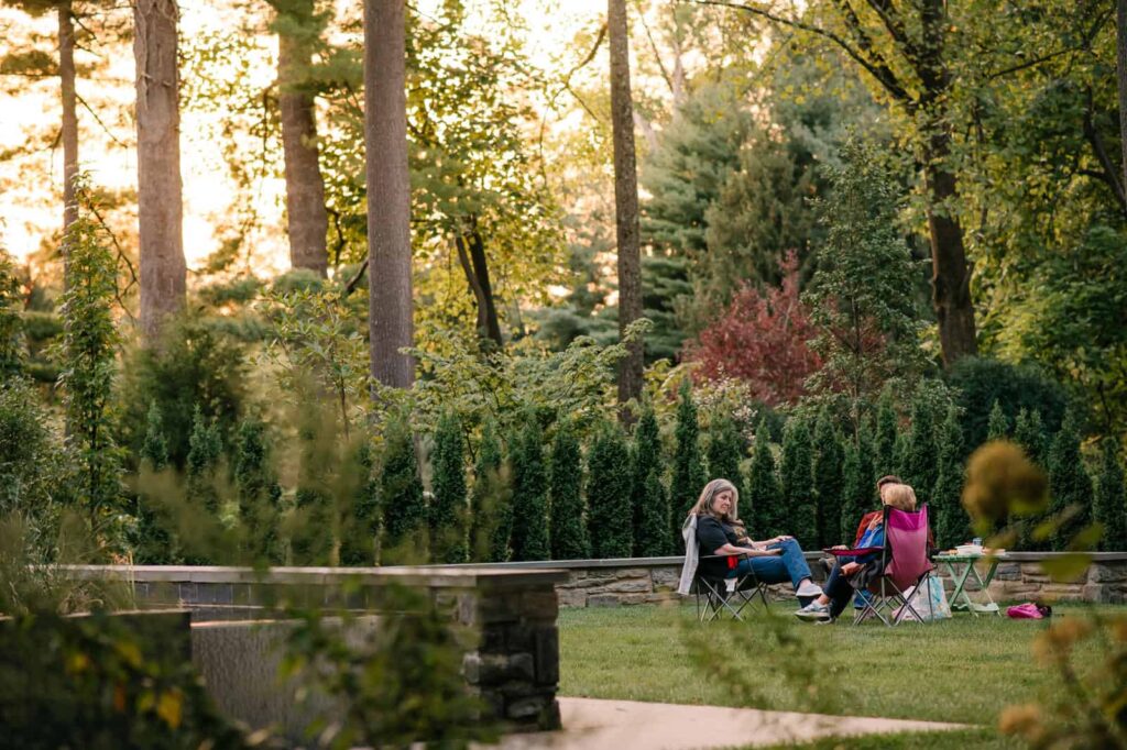 A few people sit out on the lawn as the sun sets in the distance between the trees.