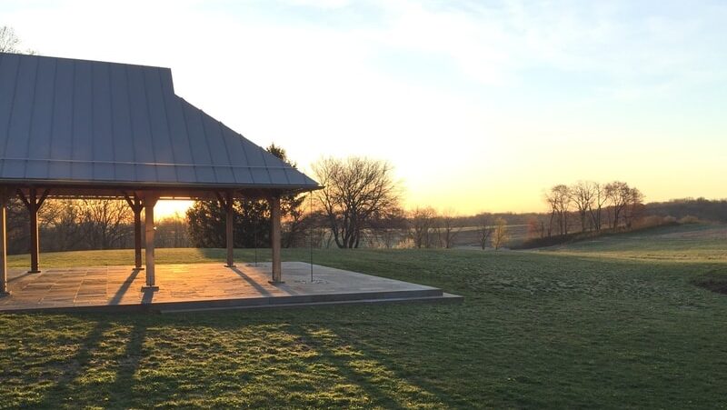 a pavilion sits in a large green field with its shadow cast on the grass as the sunrise shines behind it.