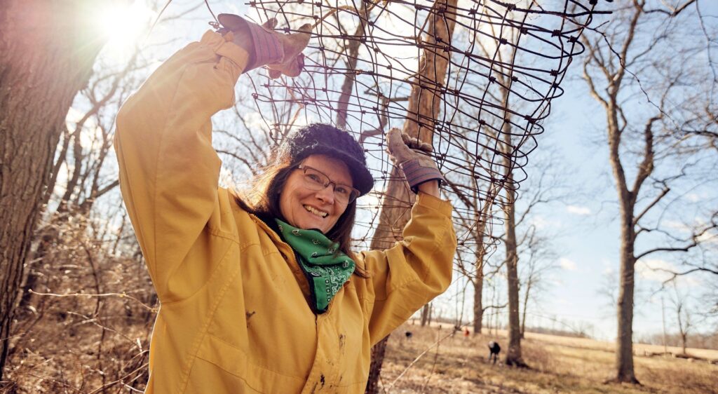 A person in a yellow jacket smiles and holds some plant fencing above their head with gloved hands as they stand in the forest.