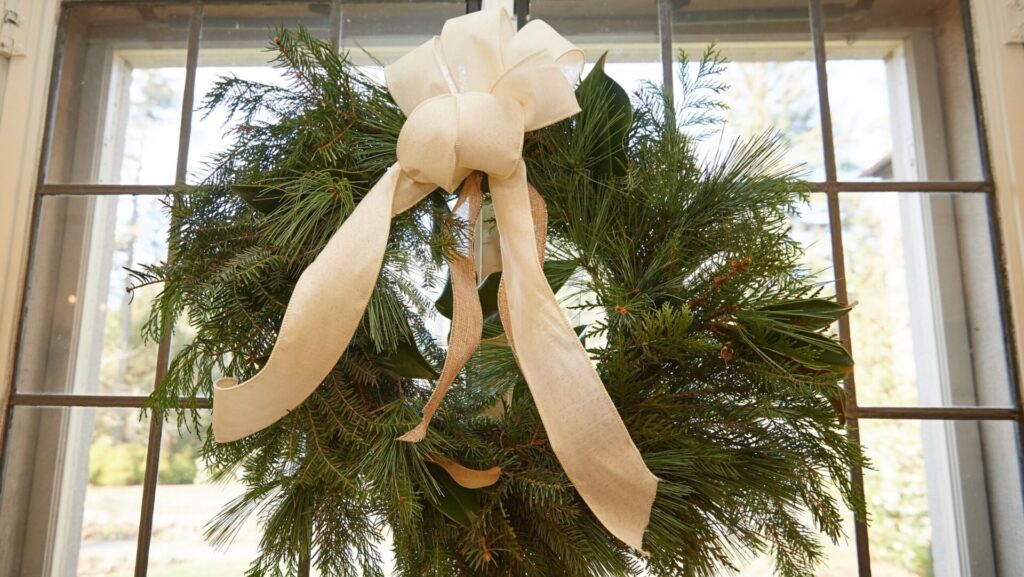 An evergreen wreath with a white bow hangs on a window.