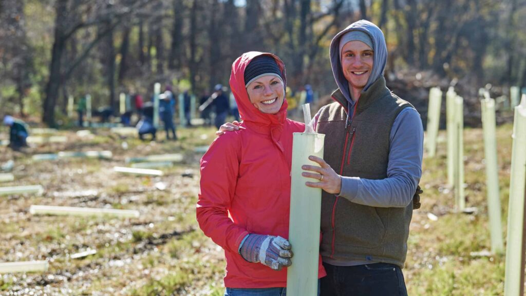 A woman with a red hooded puffy jacket and a man in a hoodie and vest smile as they place a tree tube in a field.