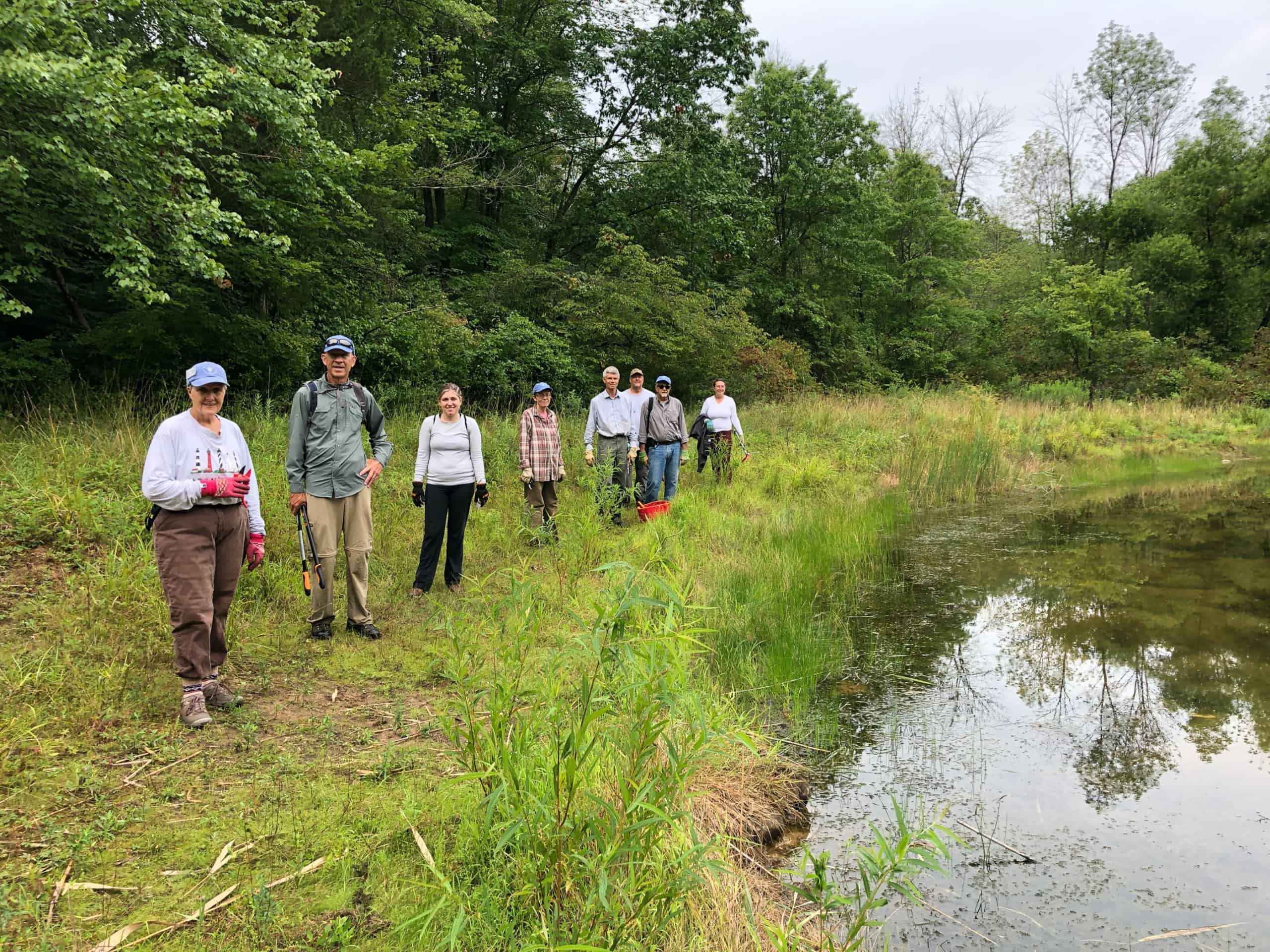 a group of people stand outside in the woods by a pond