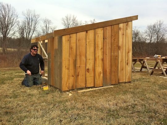 man sits next to a finished wooden shed he built