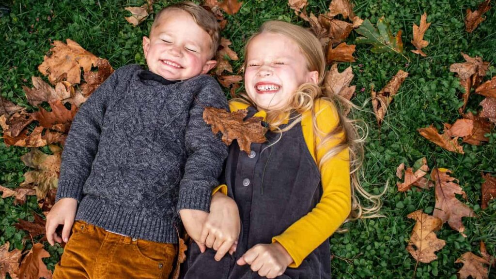 A girl and a boy in fall clothing smile with their eyes closed while lying on green grass and brown autumn leaves.