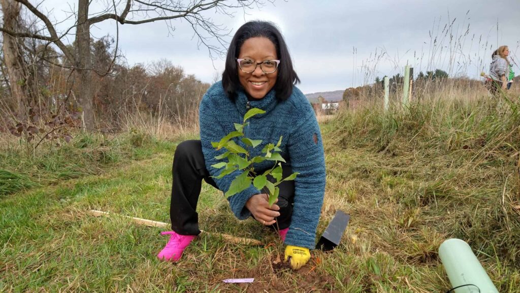 Person with glasses smiling and planting a tree sapling into the ground