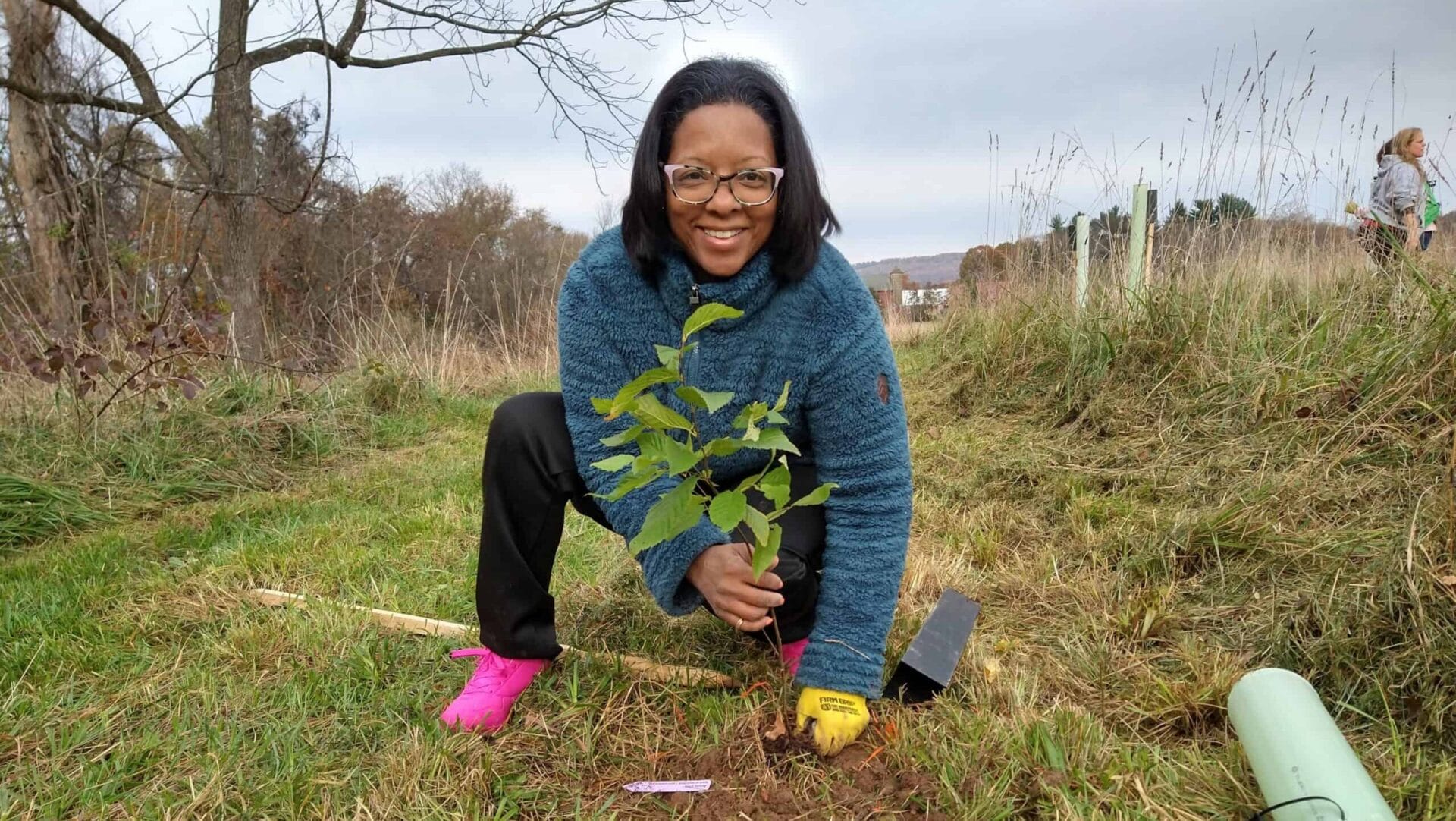 Person with glasses smiling and planting a tree sapling into the ground