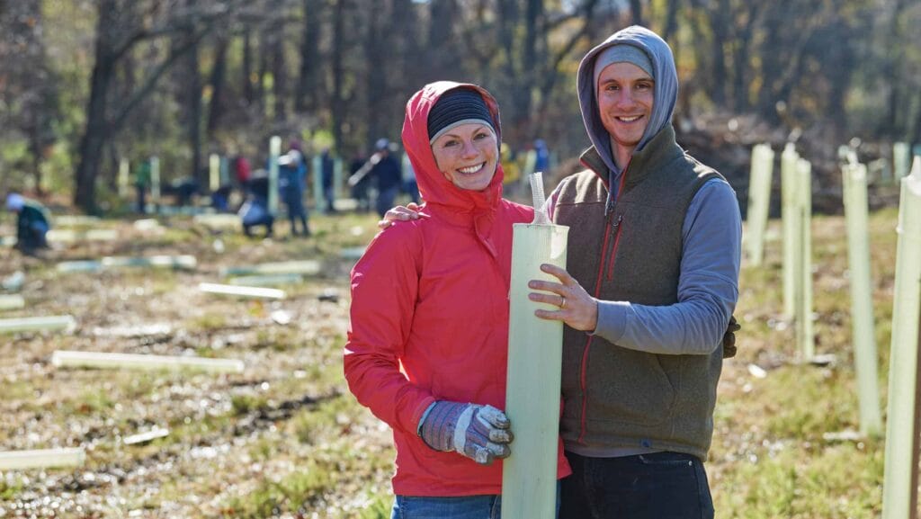 Two volunteers posing with a tree tube with many tree tubes behind them