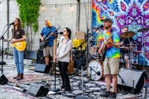 a band performing on stage with three singers, two musicians in the back and a tie dye tapestry behind