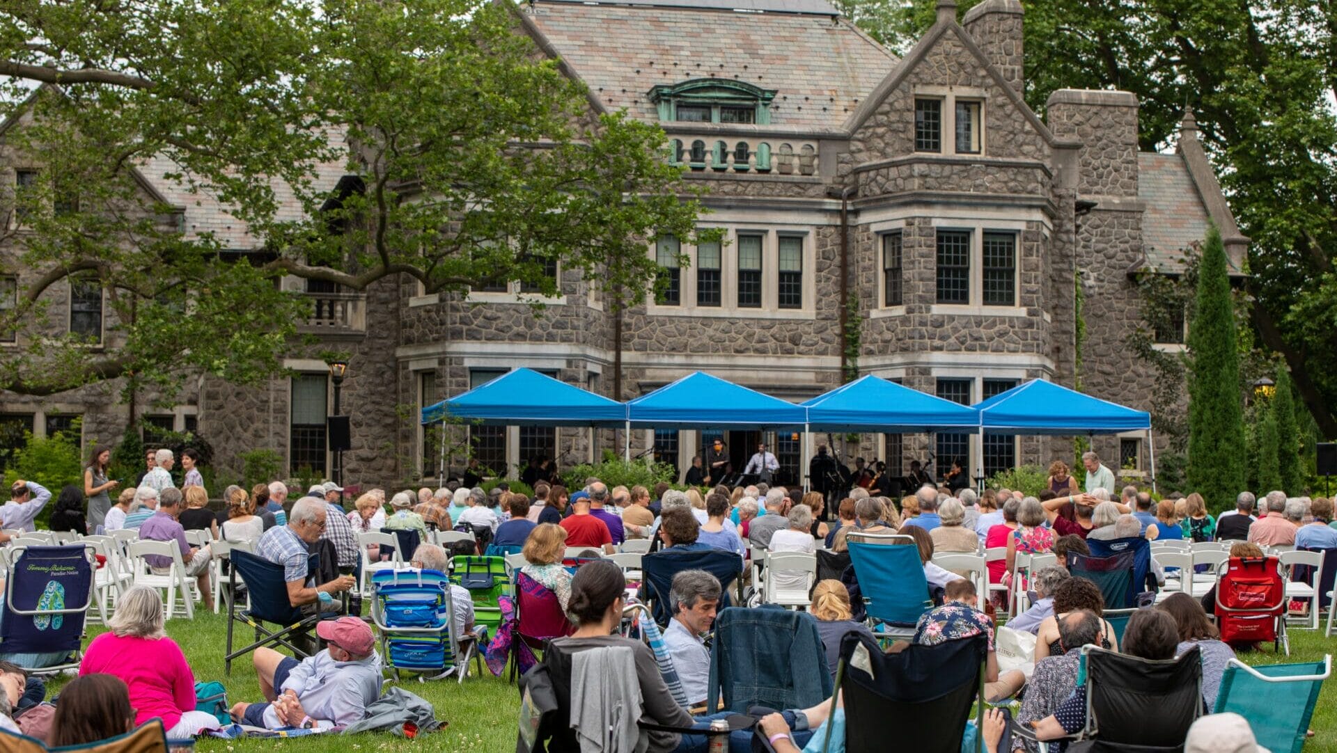a crowd of people in a lawn on blankets and chairs in front of an orchestra under tents preforming in front of a stone mansion