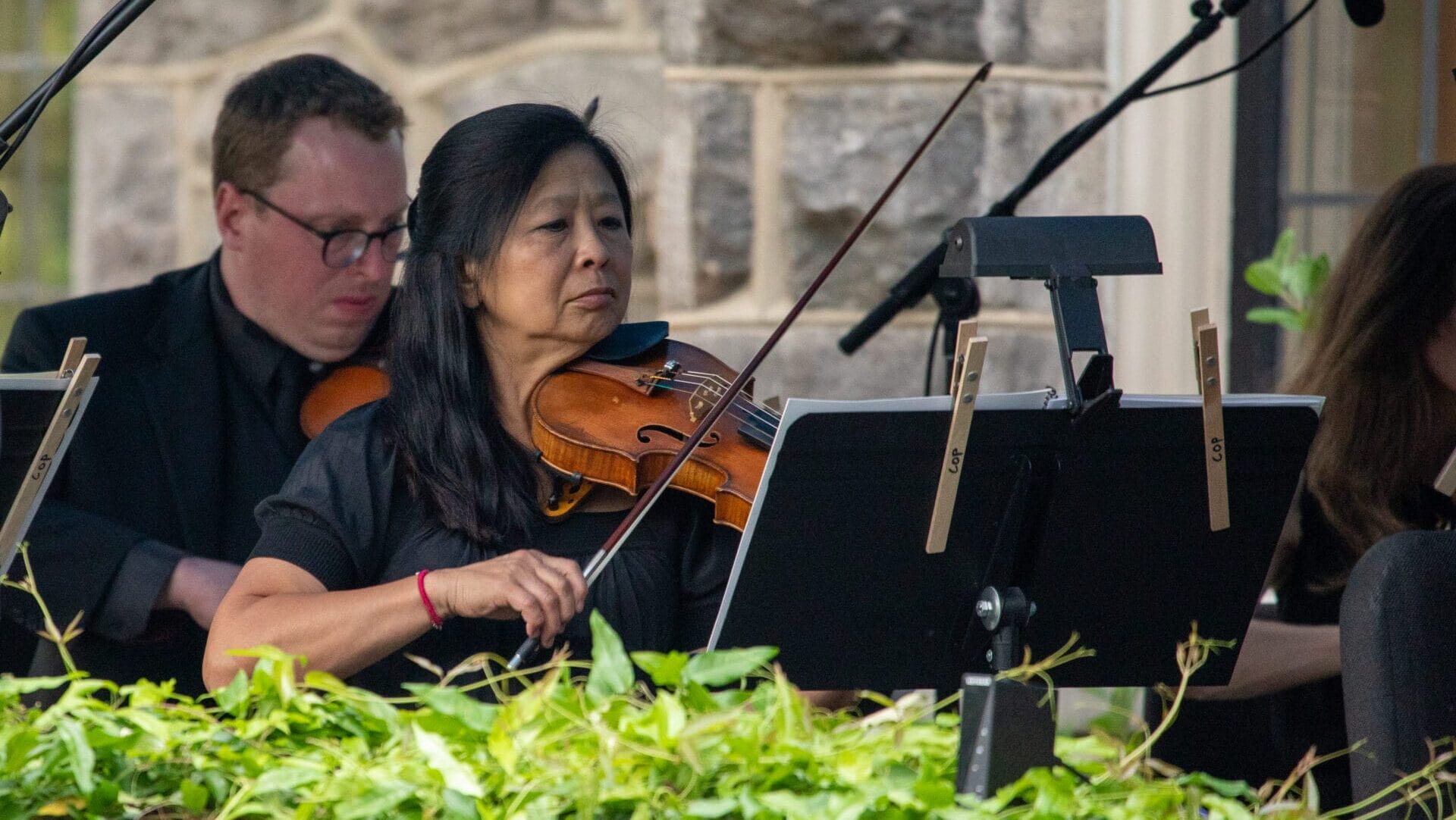 people playing violins with green leaves in the foreground and stone mansion in the background