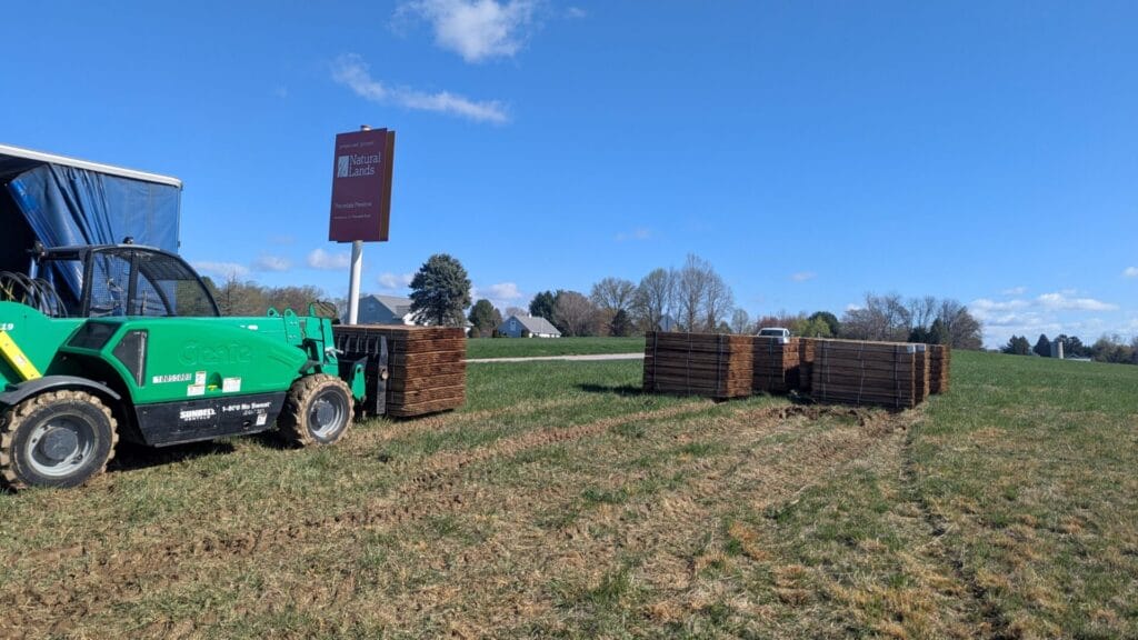 A green telehandler with pallet forks moves pallets of stakes onto a nature preserve.