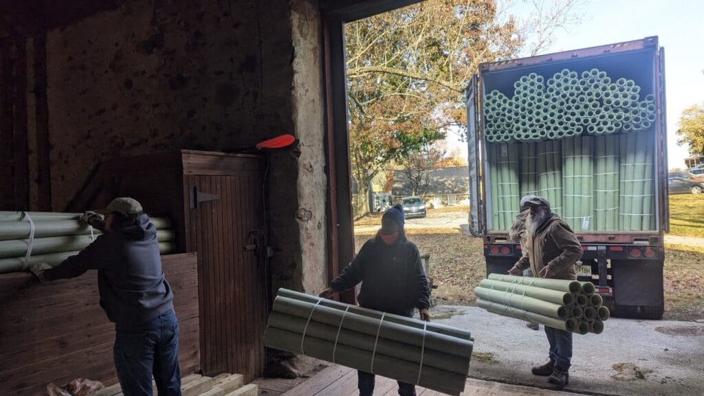 Three people unload bundles of protective plastic tree tubes from a truck into a barn.