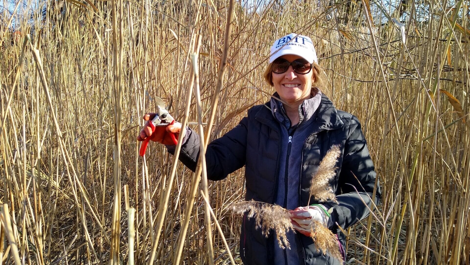a smiling woman with sunglasses, hat, and coat pruning tall grasses