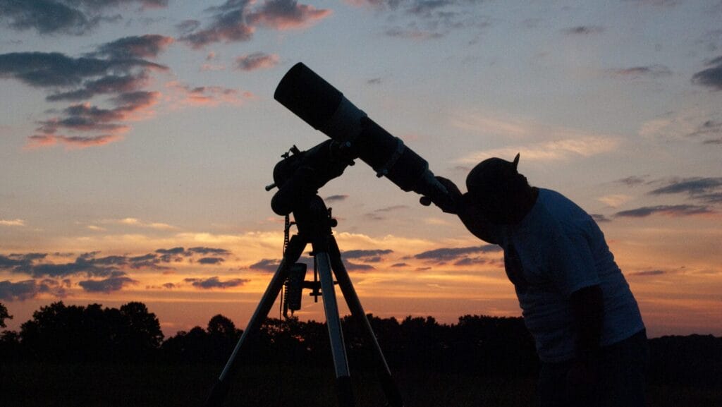 The silhouette of a person looking up through a telescope with a vibrant orange and blue sunset in the background.