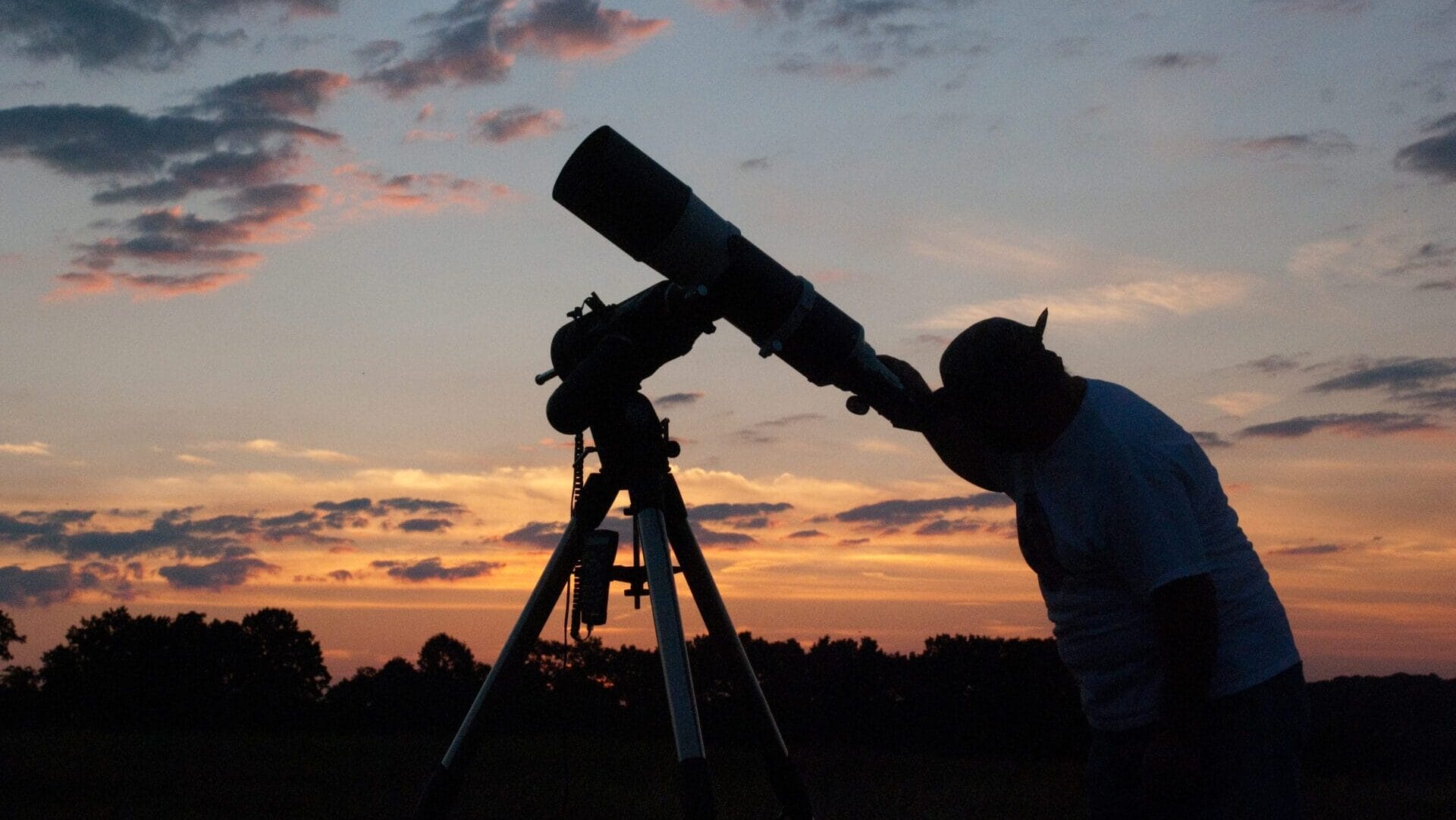 The silhouette of a person looking up through a telescope with a vibrant orange and blue sunset in the background.