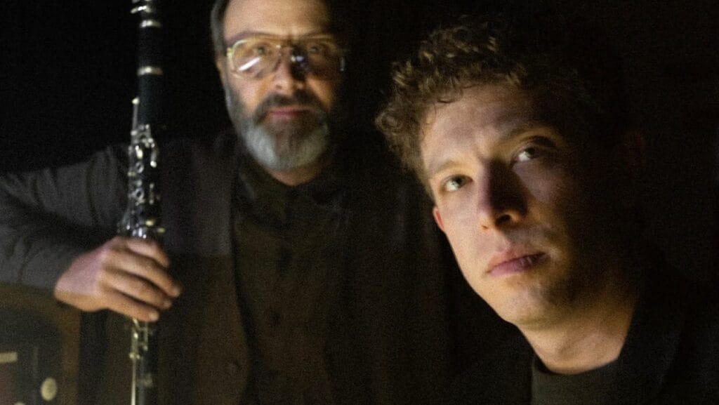 two men wearing black in front of a black background, one holding a clarinet