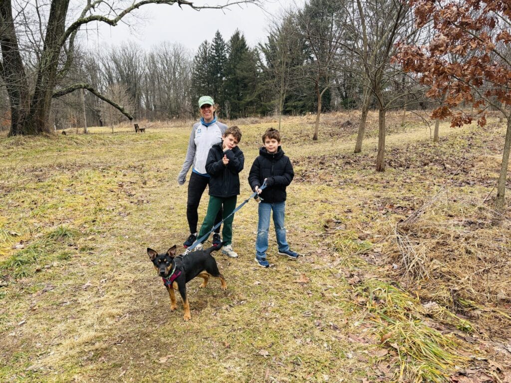 A family of three and a medium-size black and tan dog on a leash walk a grassy path outdoors