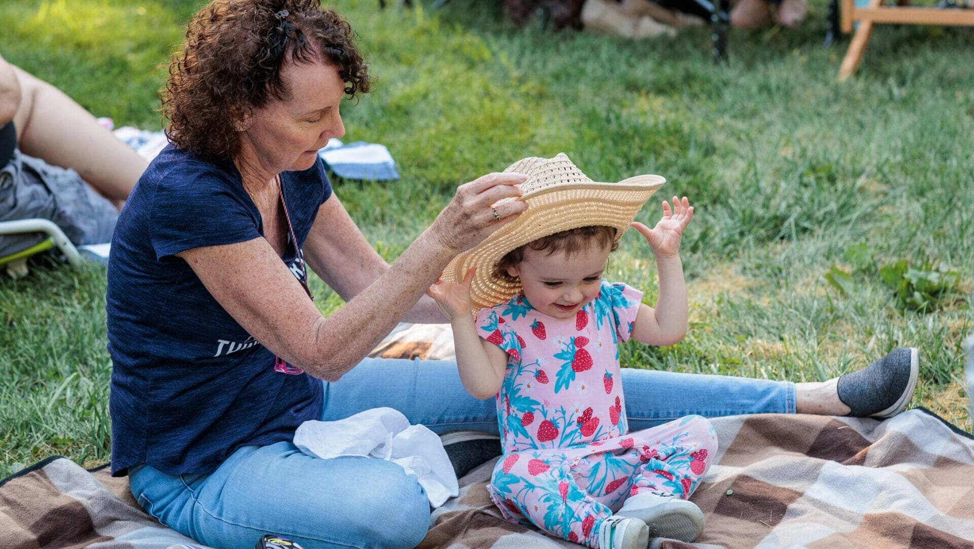 A woman placing a straw hat on a child's head while sitting on a blanket outside.