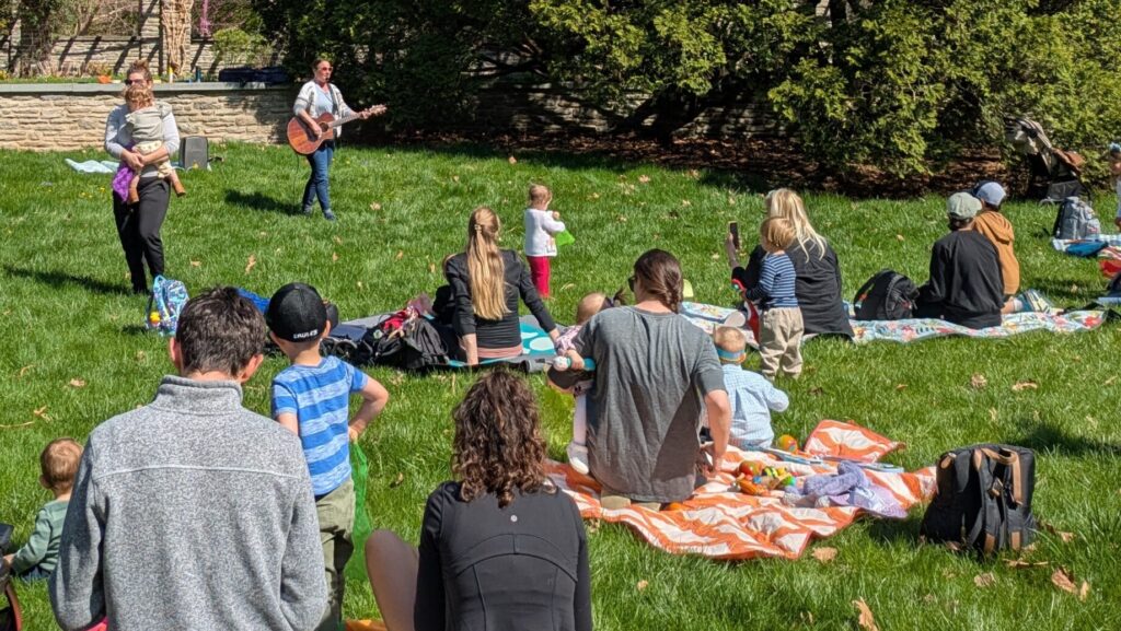 People and children on blankets on the lawn with a woman playing guitar.