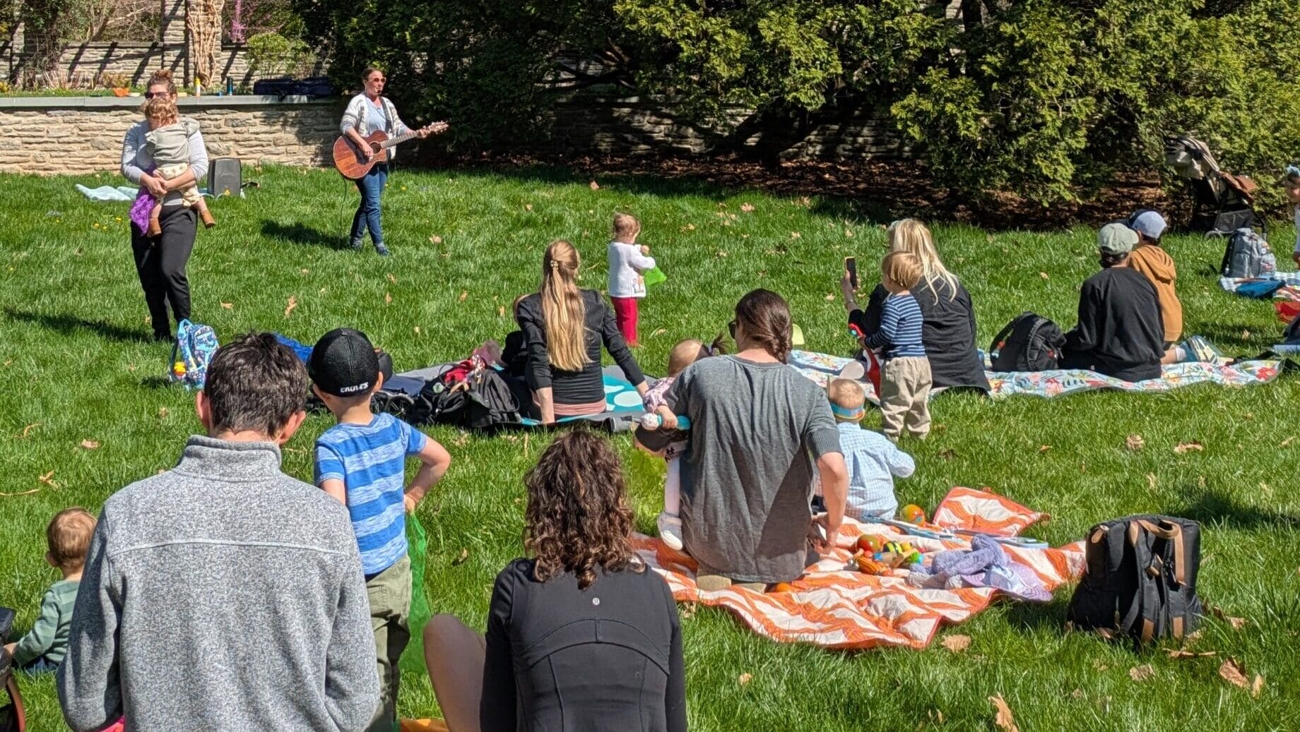 People and children on blankets on the lawn with a woman playing guitar.