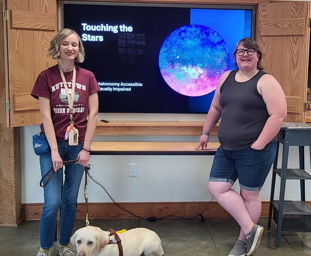 Two women stand in front of a presentation screen. One has a seeing eye dog