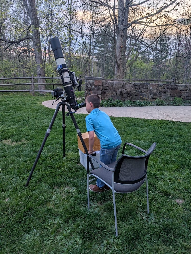 A boy in a blue shirt with his eye up to the viewer of a large telescope set in a grassy nature area.